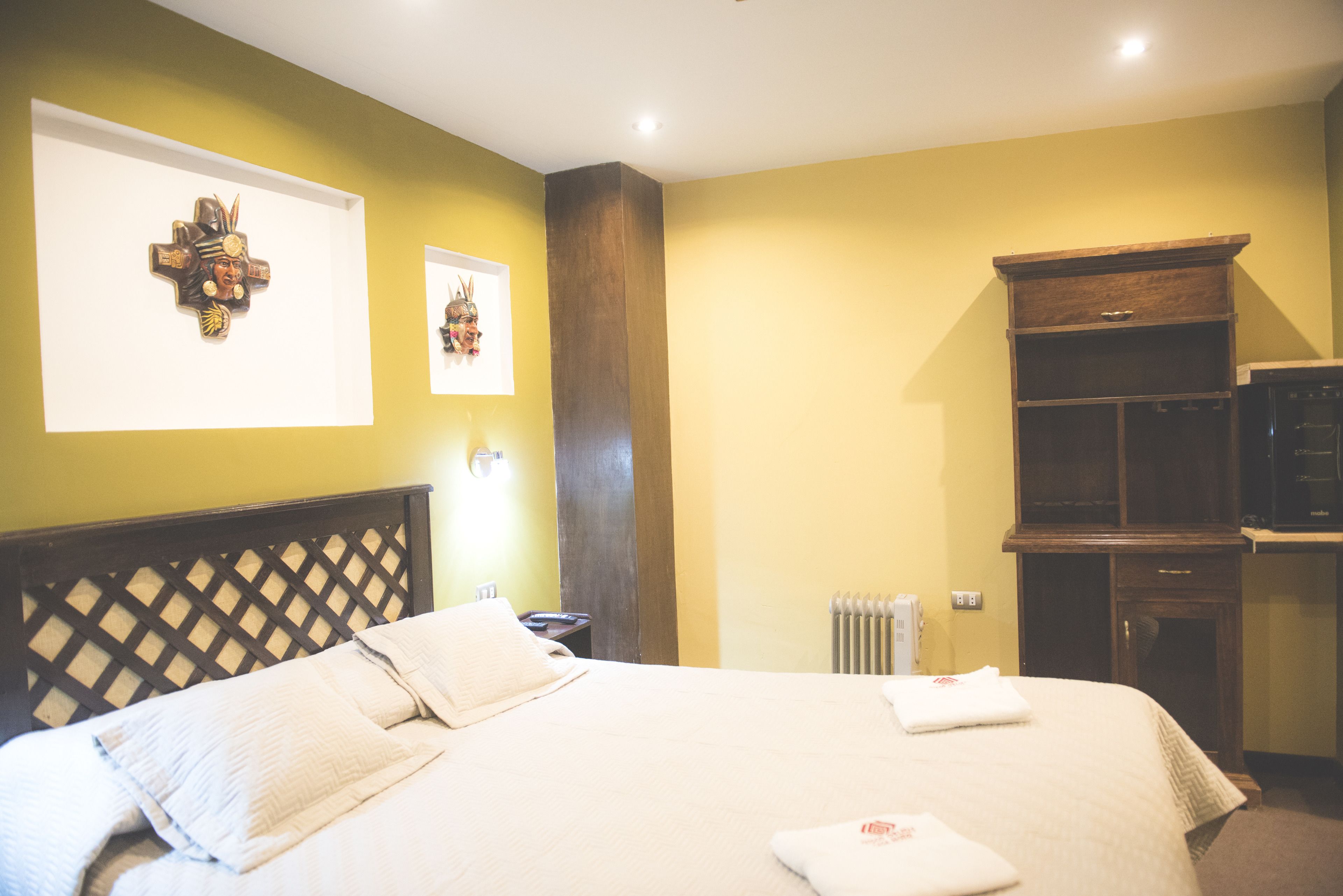 A hotel room with a large bed, a wooden headboard, two wall-mounted artworks, a built-in TV cabinet, and a radiator.