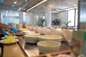 Montaje de buffet con múltiples fuentes y platos de comida, una persona sirviendo, y un interior moderno con separaciones de vidrio.