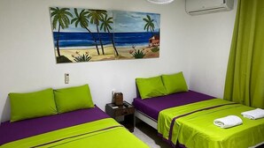 Ett hotellrum med två sängar, var och en med grönt och lila sängkläder, en tavla med ett strandmotiv på väggen och en grön gardin.