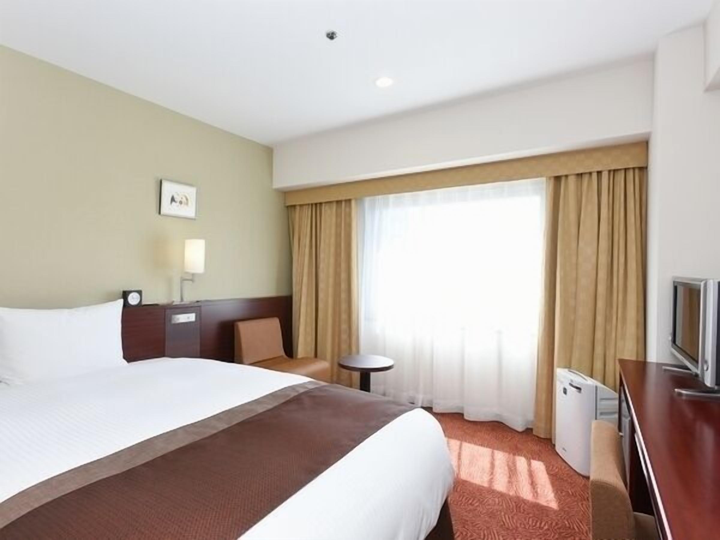 Ein Hotelzimmer mit einem großen Bett, einem Schreibtisch mit Fernseher, einem kleinen Tisch und einem Fenster mit Vorhängen.