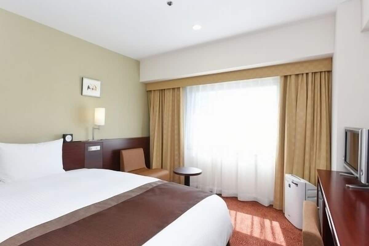 A hotel room with a large bed, a desk with a TV, a small table, and a window with curtains.