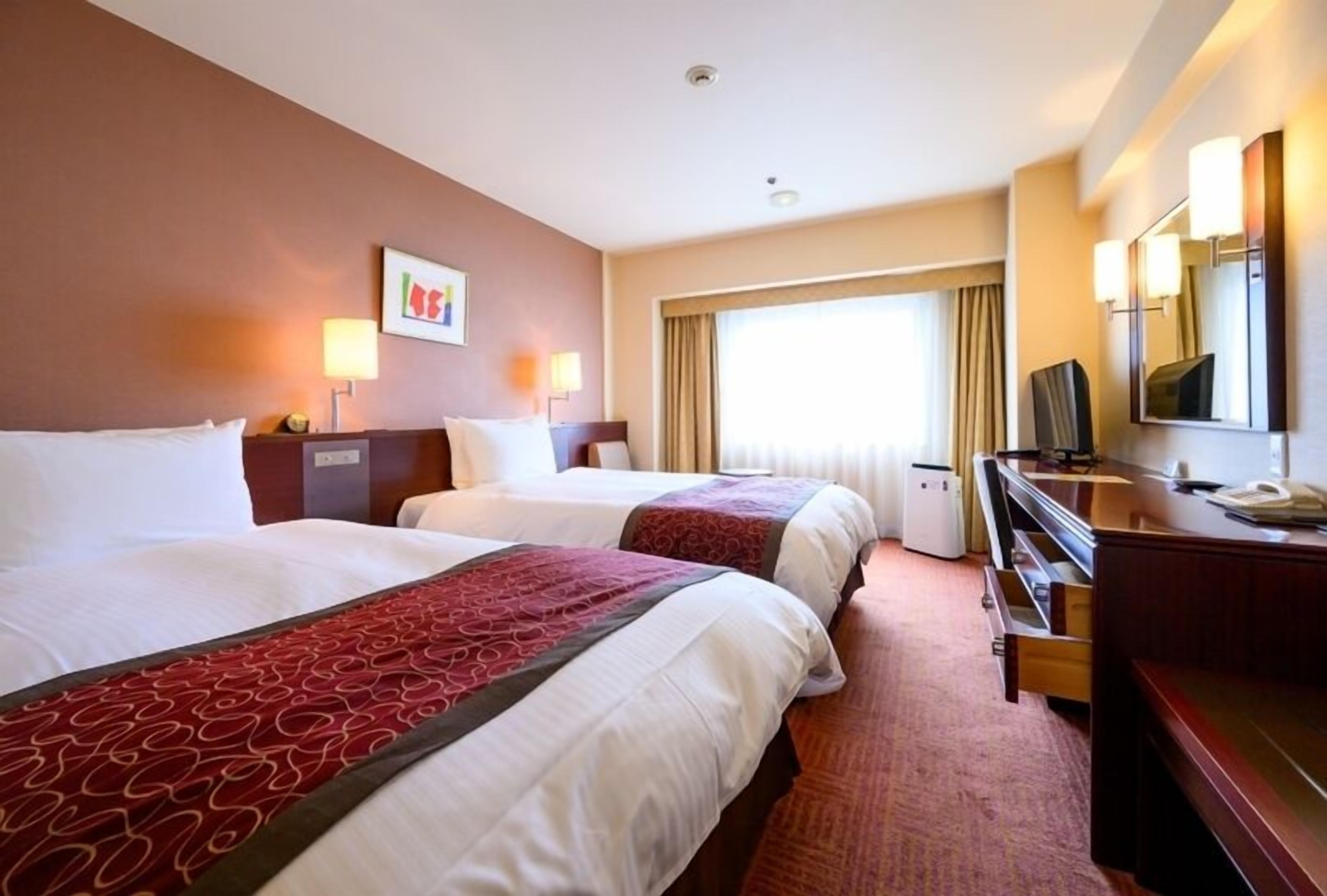 Ein Hotelzimmer mit zwei Betten, einem Schreibtisch, einem Fernseher und einem großen Fenster mit Vorhängen.