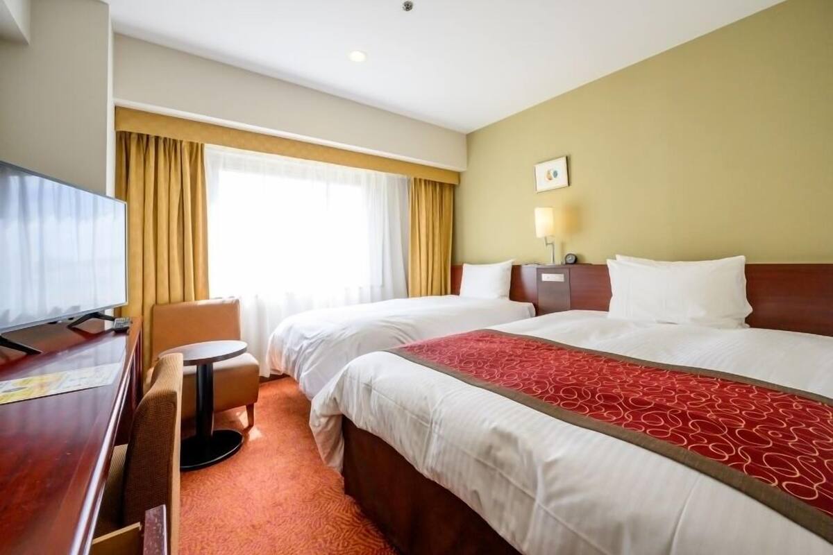 A hotel room with two beds, a desk, a chair, a TV, and a window with curtains.