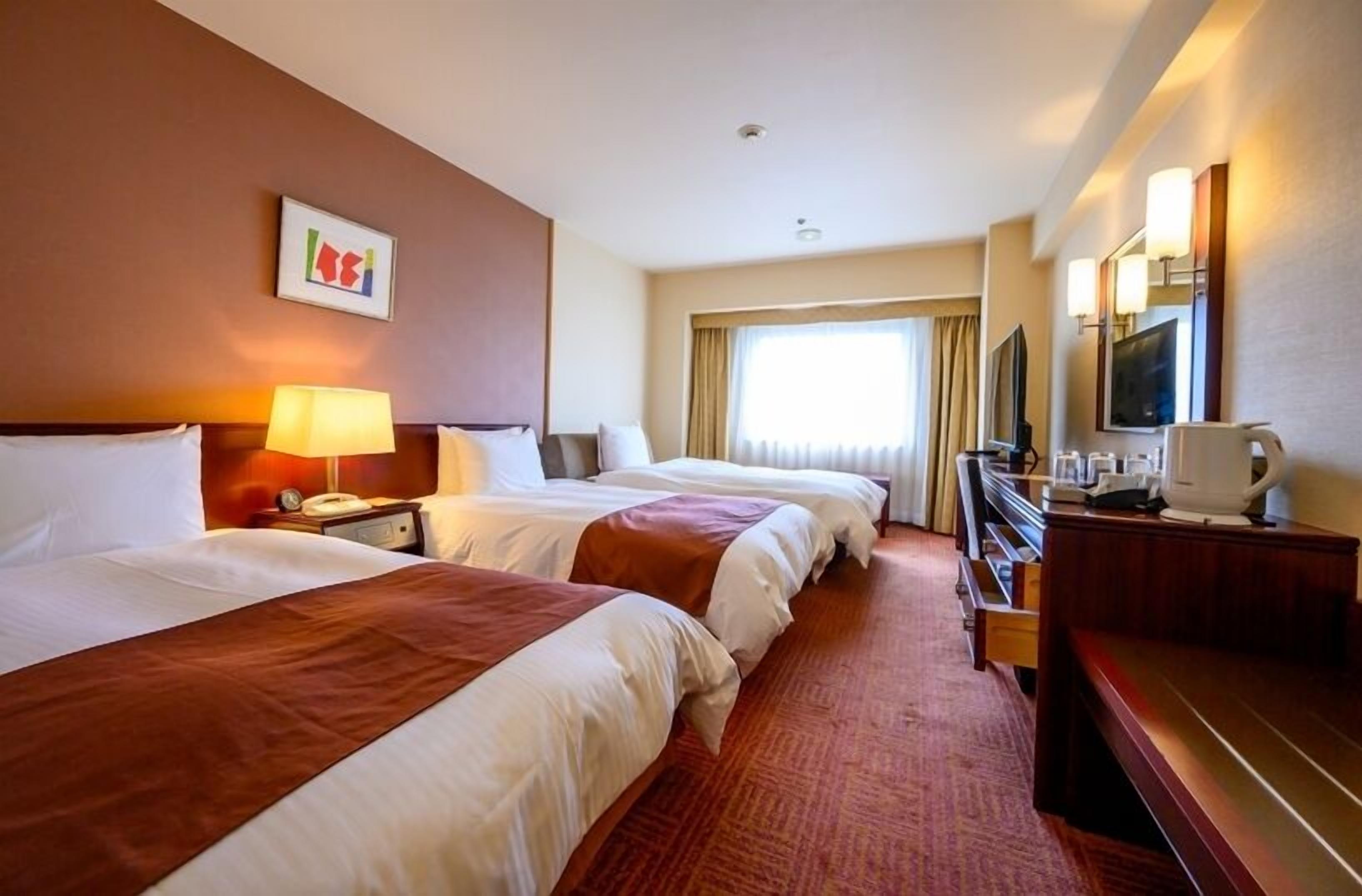 Ein Hotelzimmer mit zwei Betten, einem Schreibtisch mit Computer, einem Fernseher und einem Fenster mit Vorhängen.
