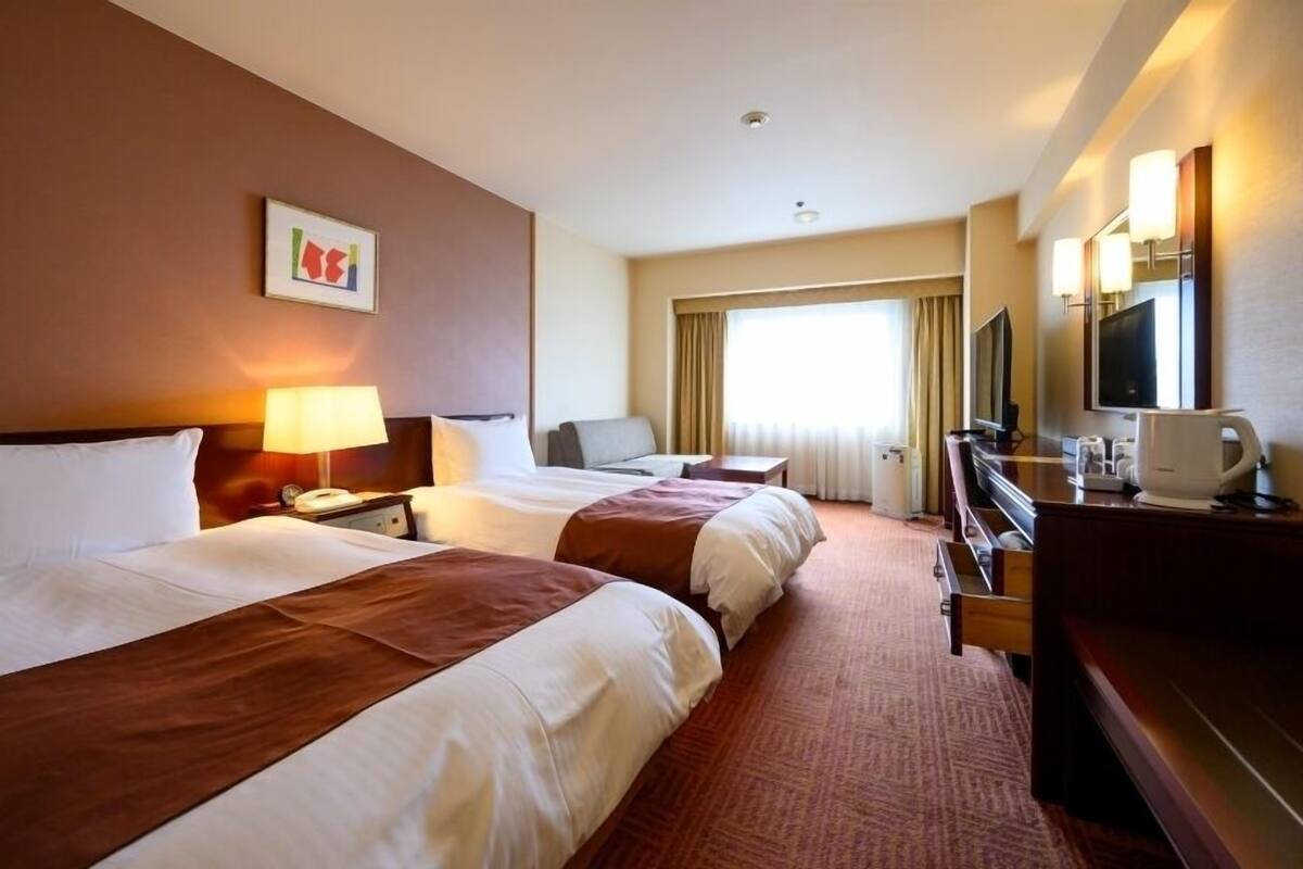 A hotel room with two beds, a desk, a chair, a television, and a window with curtains.