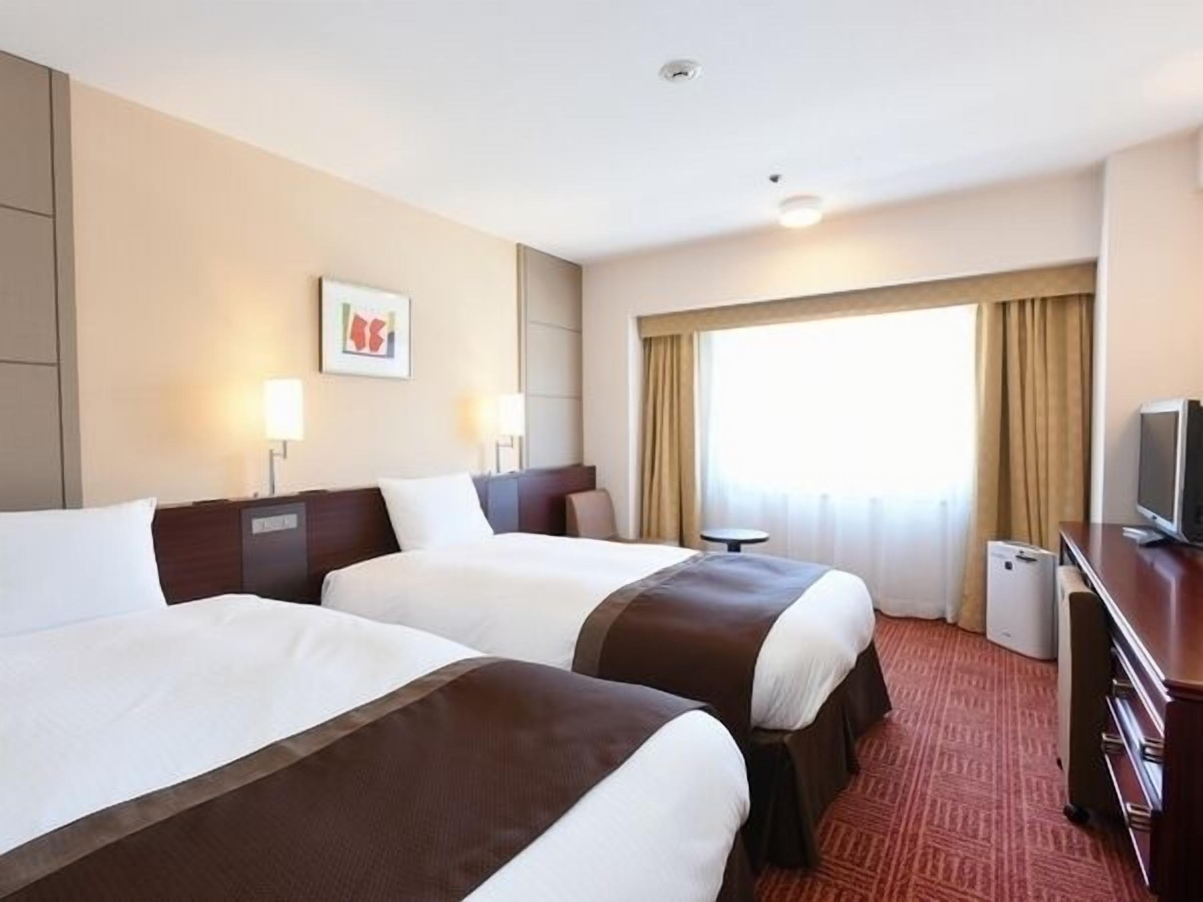 Ein Hotelzimmer mit zwei Betten, einem Schreibtisch, einem Fernseher und einem Fenster mit Vorhängen.