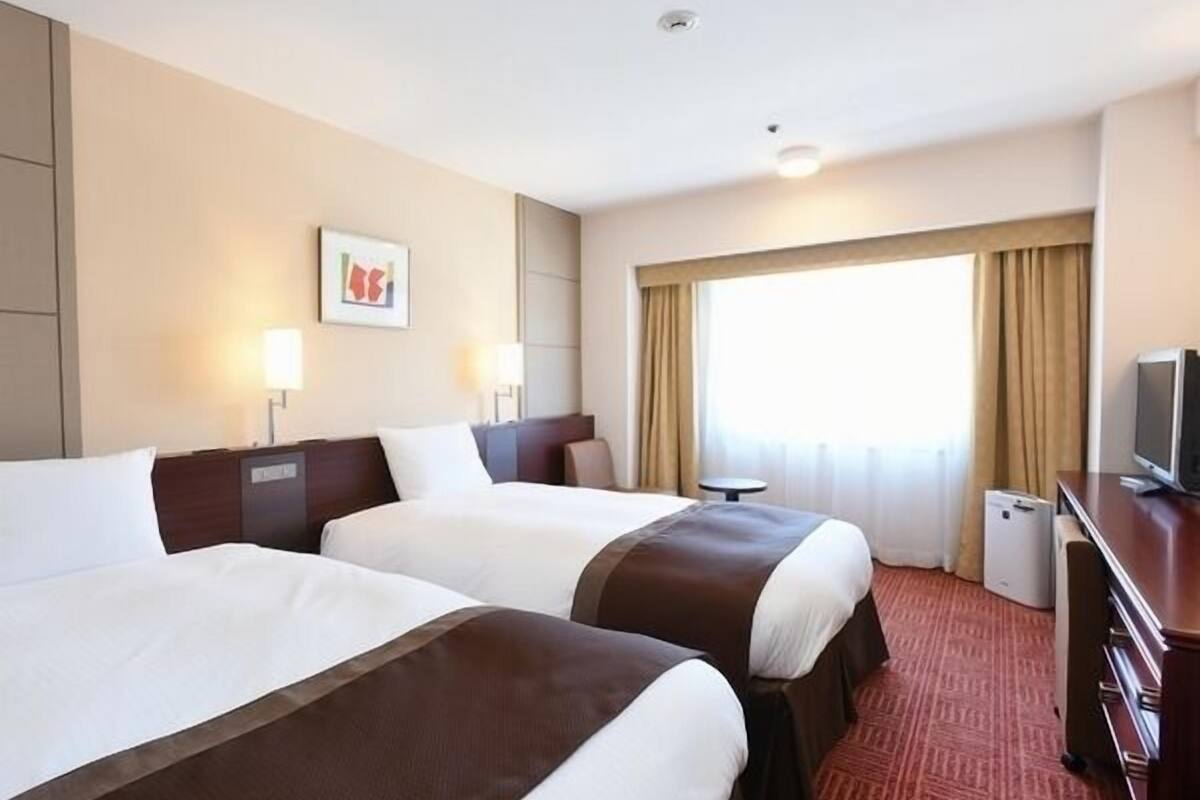 A hotel room with two beds, a desk, a television, and a window with curtains.