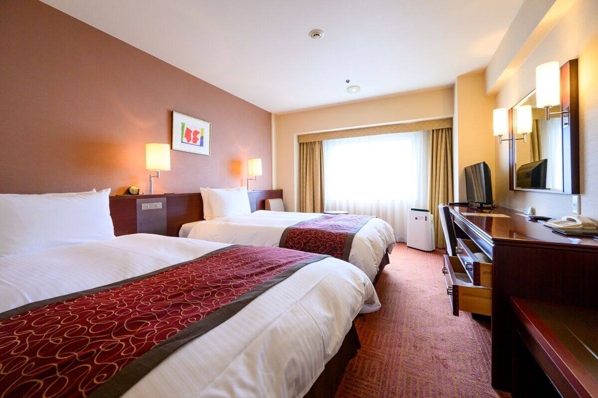 A hotel room with two beds, a desk, a television, and a large window with curtains.