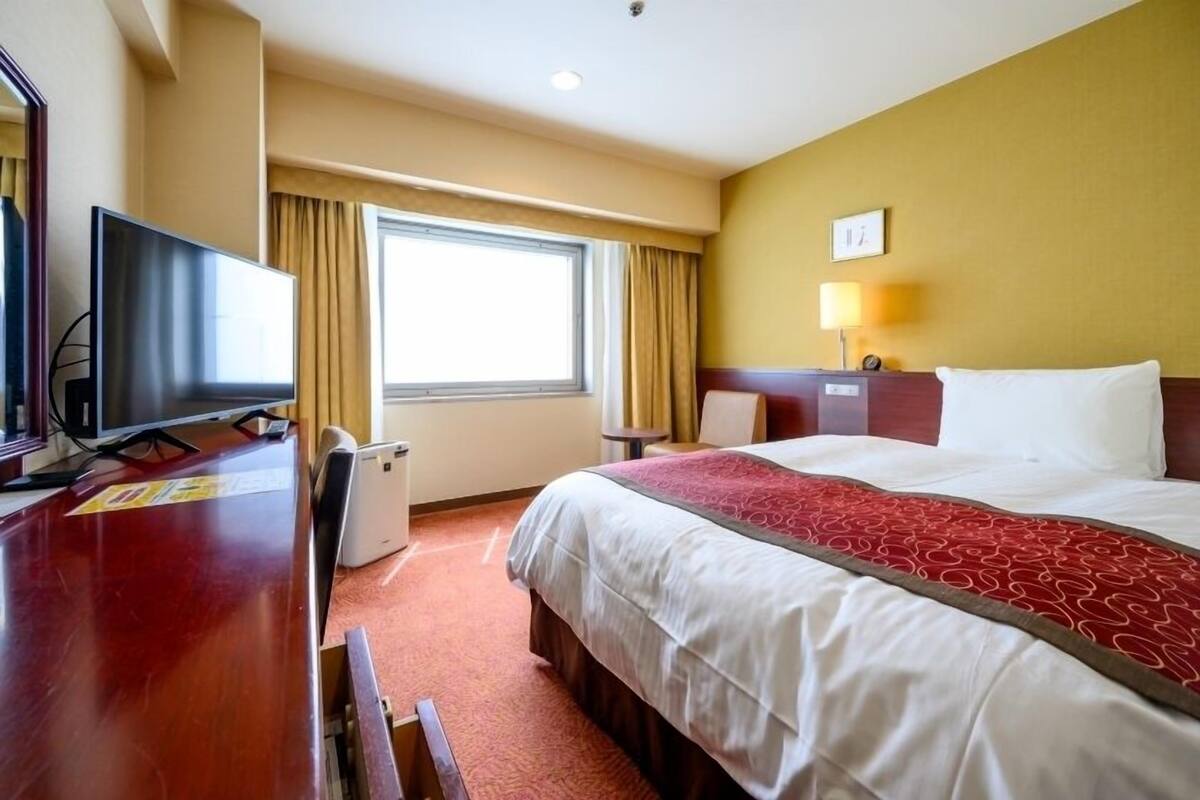 A hotel room with a bed, a desk with a TV, a window with curtains, and a bedside lamp.