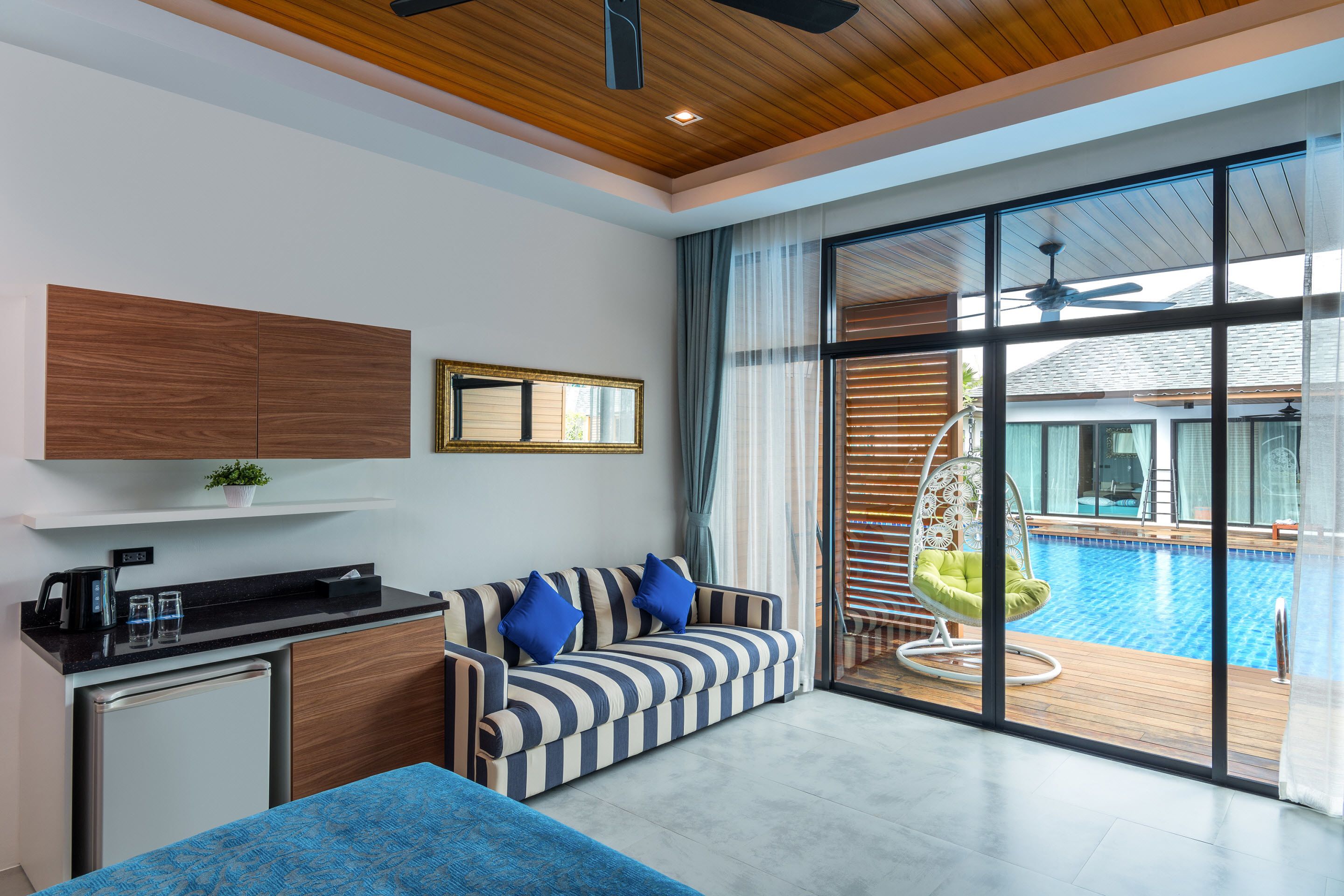 Una habitación moderna con sofá, kitchenette pequeño y amplio ventanal con vista a una piscina.