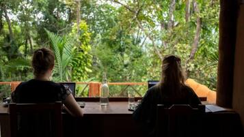 Two individuals seated at a wooden table with laptops, overlooking a lush green forest.