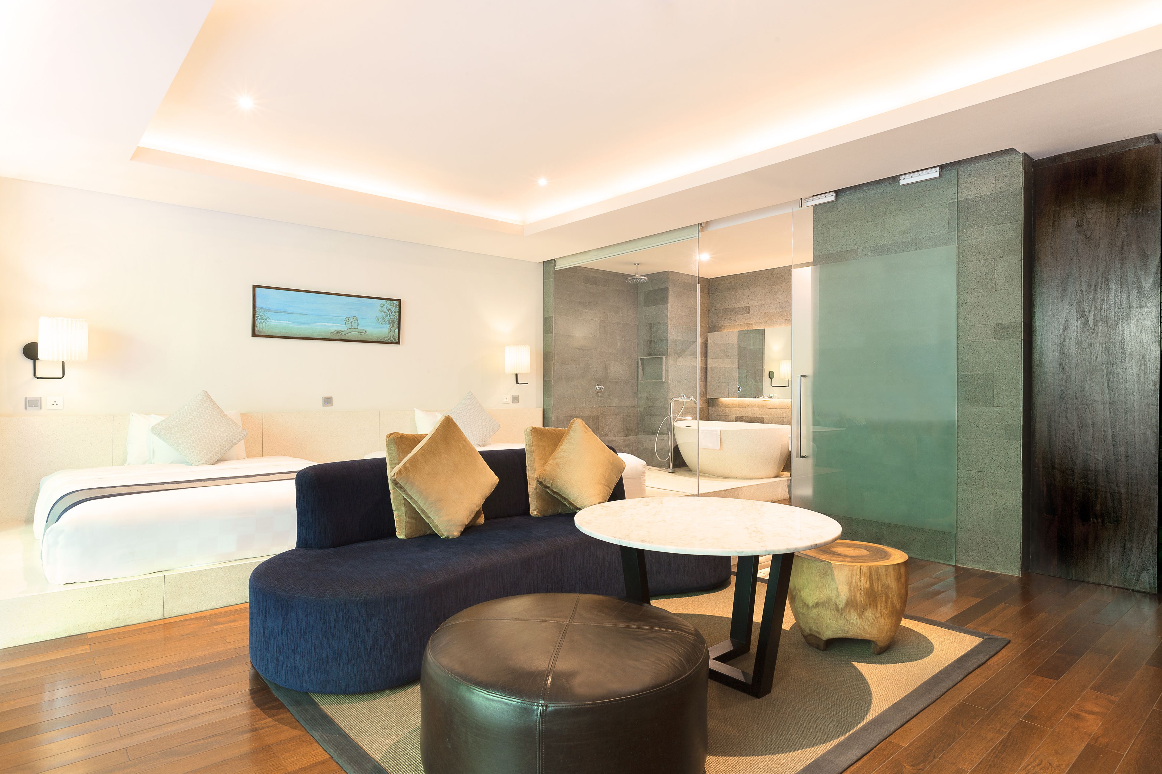 Ein modernes Hotelzimmer mit Sofa, rundem Tisch, Holzschemel, Bett und einer Duschkabine aus Glas.