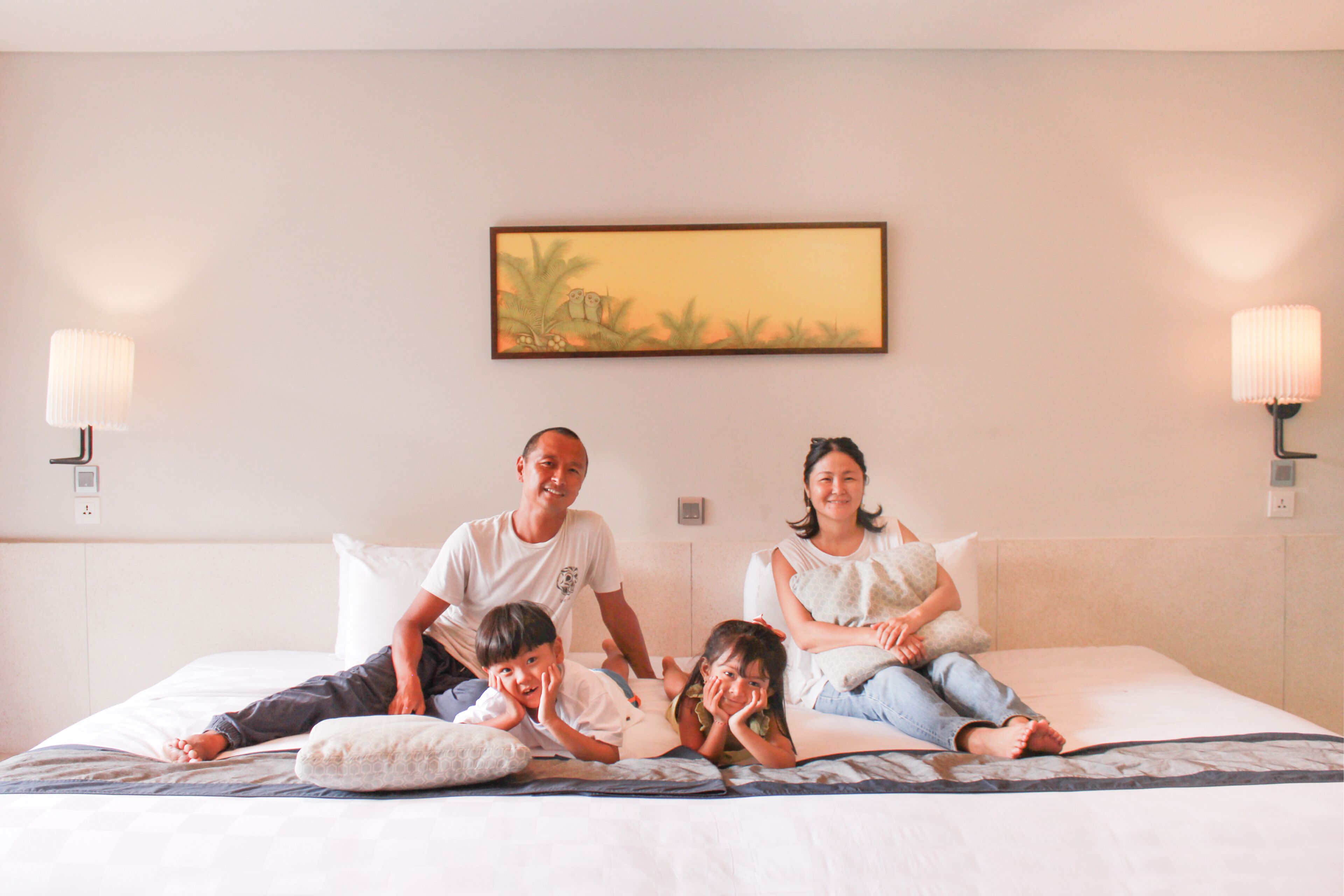 A family of four on a bed with a painting above them and wall sconces on either side.
