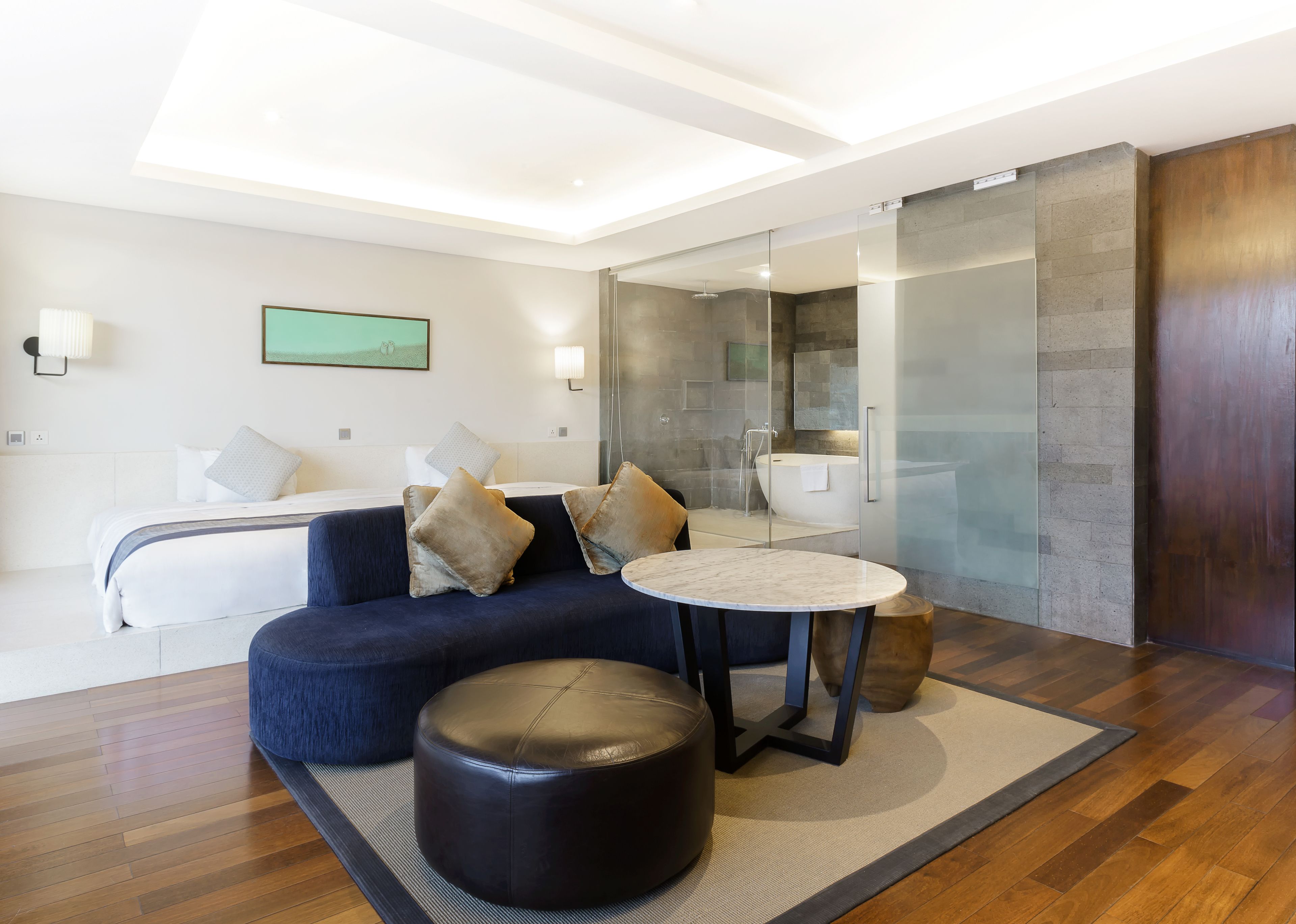 Quarto de hotel moderno com cama, sofá azul, mesa redonda e banheiro com paredes de vidro.