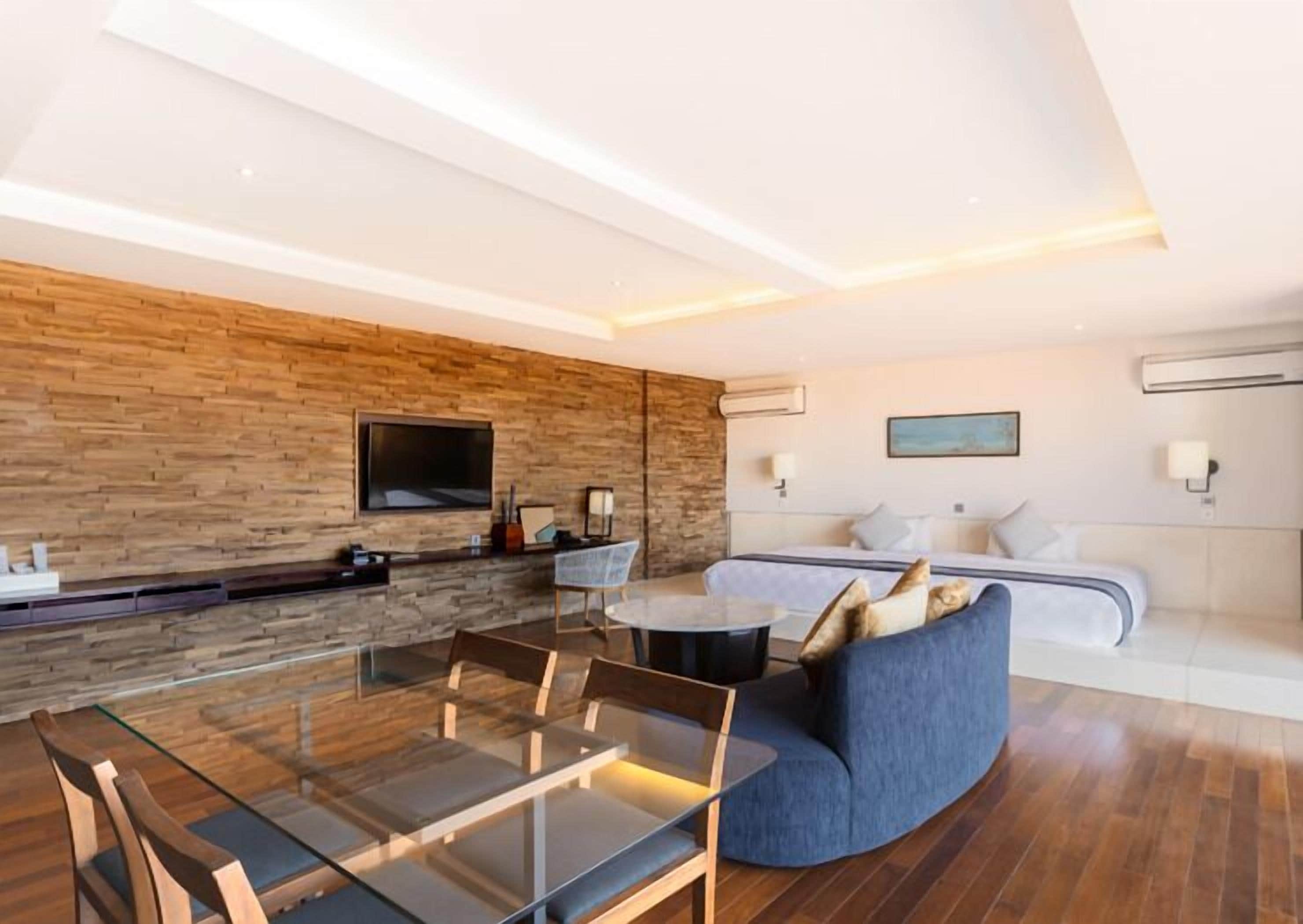 Een moderne woonkamer met een glazen eettafel, een bank en een televisie bevestigd aan een houten wand.