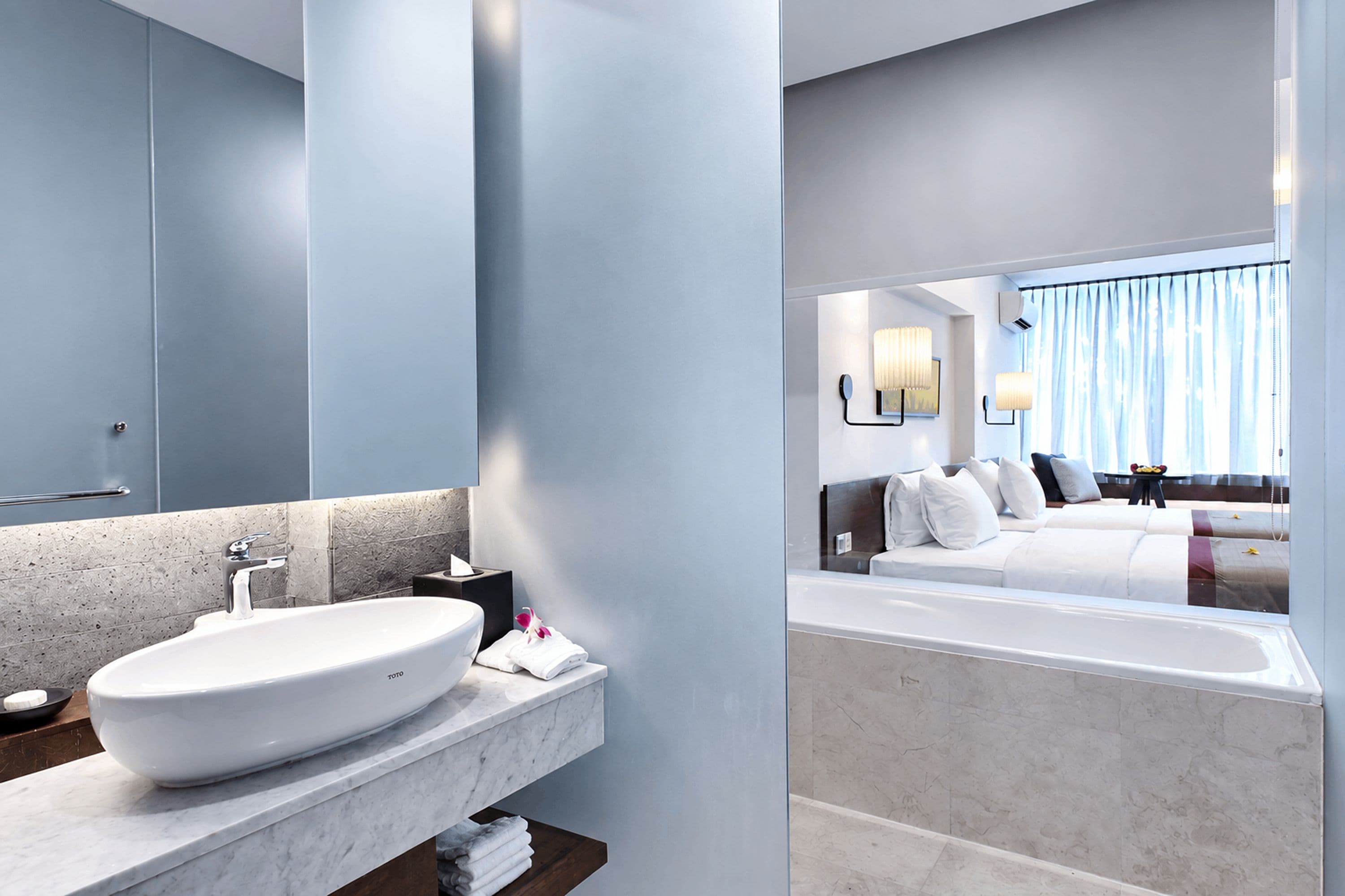 A modern bathroom with a large sink, mirror, and a view into a bedroom with a bed and curtains.