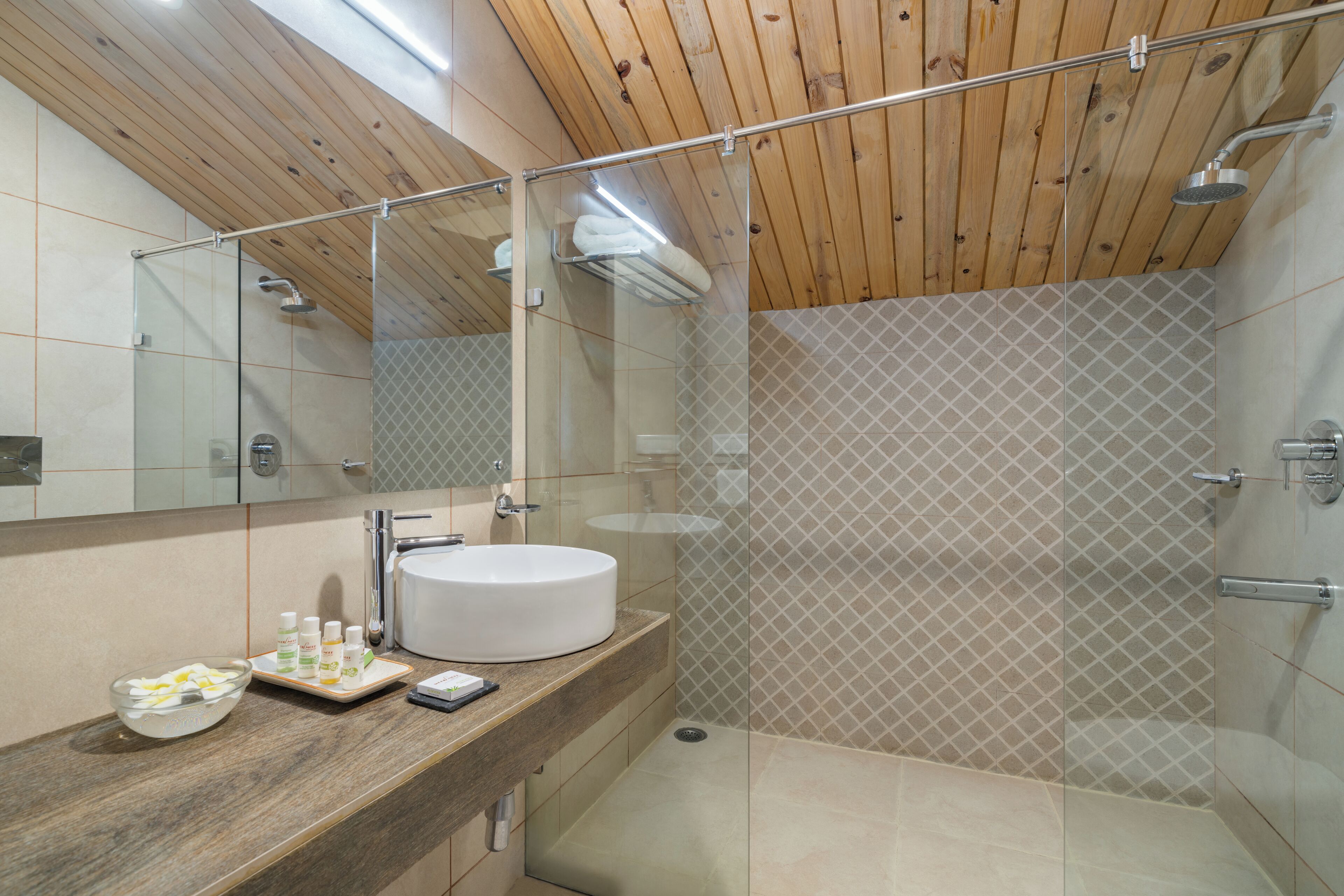 Ein modernes Badezimmer mit Holzboden, einer Dusche mit Glaswand und einem weißen Waschbecken mit Pflegeprodukten.