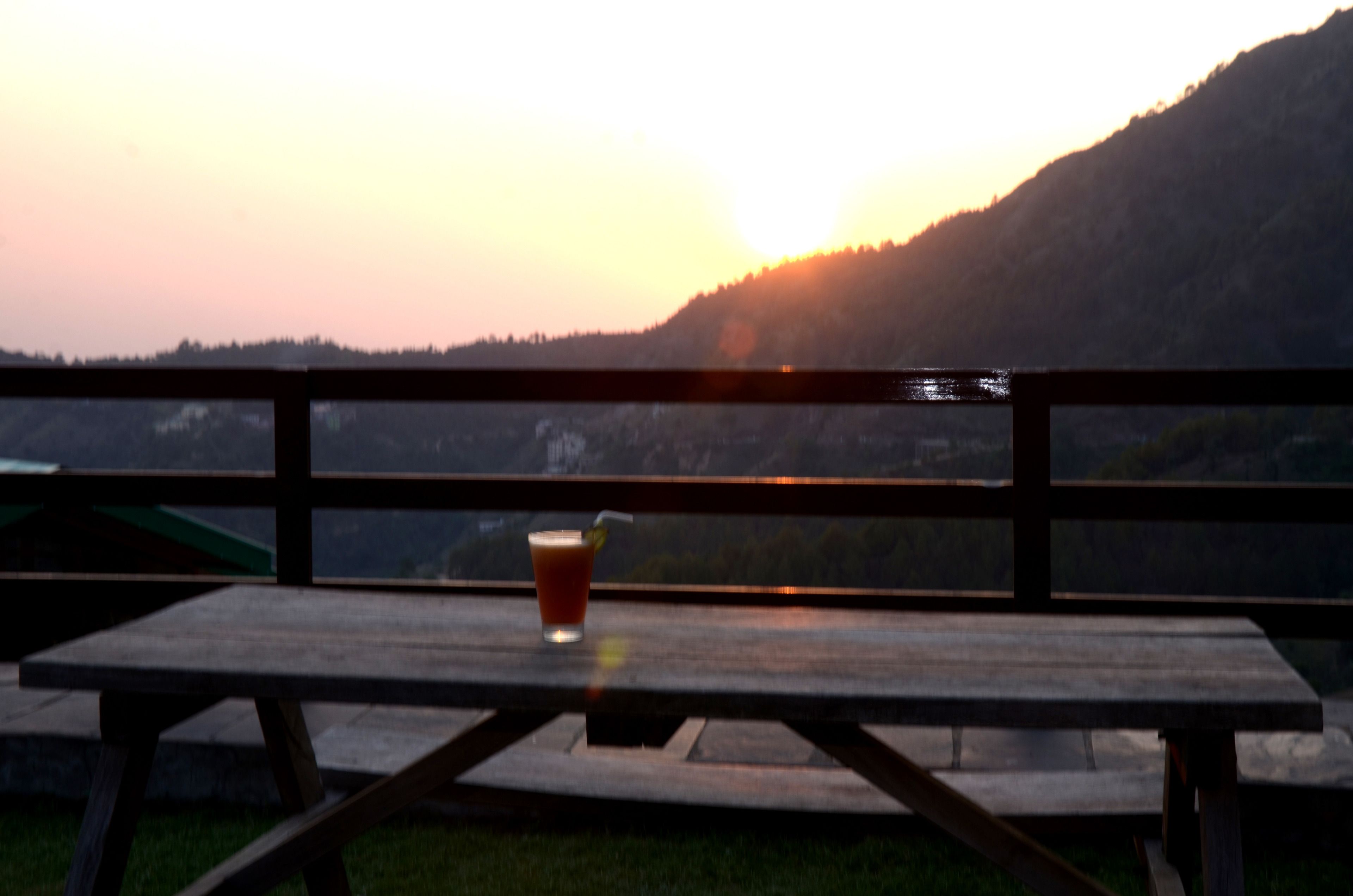 Ein Holztisch für ein Picknick mit einem Getränk und einem Strohhalm, vor einer Kulisse von Sonnenuntergang über einer Berglandschaft.