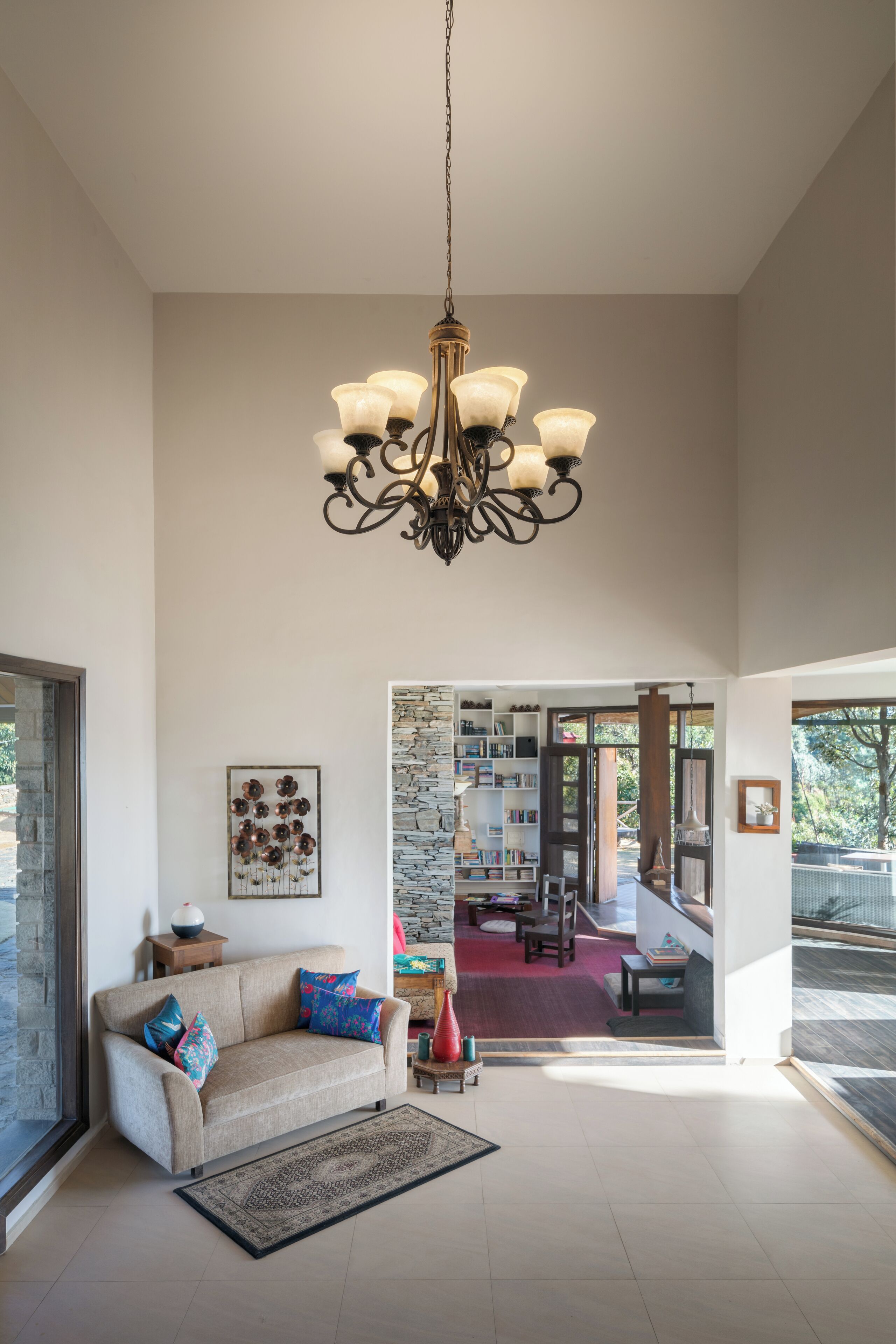 A spacious living room with a high ceiling, a chandelier, a sofa with colorful cushions, a patterned rug, and a view into a dining area with a stone wall and bookshelves.