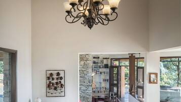 A spacious living room with a high ceiling, a chandelier, a sofa with colorful cushions, a patterned rug, and a view into a dining area with a stone wall and bookshelves.