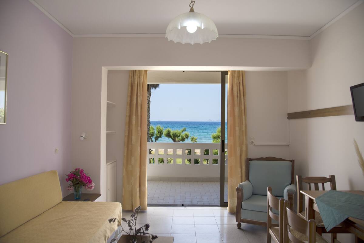 A room with a bed, a chair, a table, a TV, and a view of the sea through a balcony.