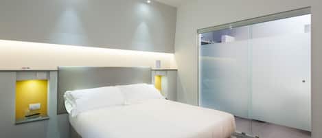Una habitación de hotel moderna con una cama grande, mesitas de noche, un armario y una pared divisoria de vidrio.