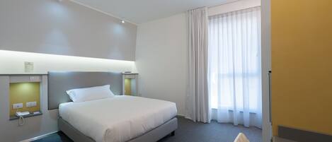 Ein Hotelzimmer mit einem großen Bett, einem Nachttisch, einem Sessel, einem Fenster mit Vorhängen und einer Tür.