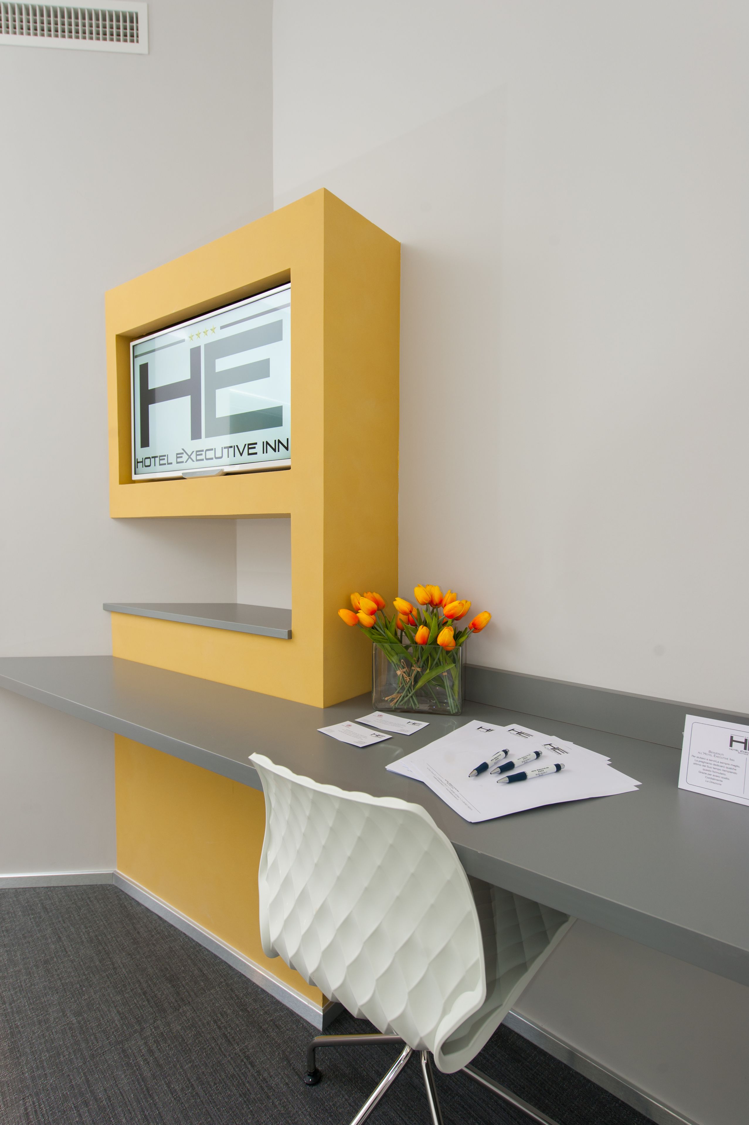 A reception desk with a yellow accent wall, a glass display case with the hotel name, a white chair, and a vase with orange flowers.
