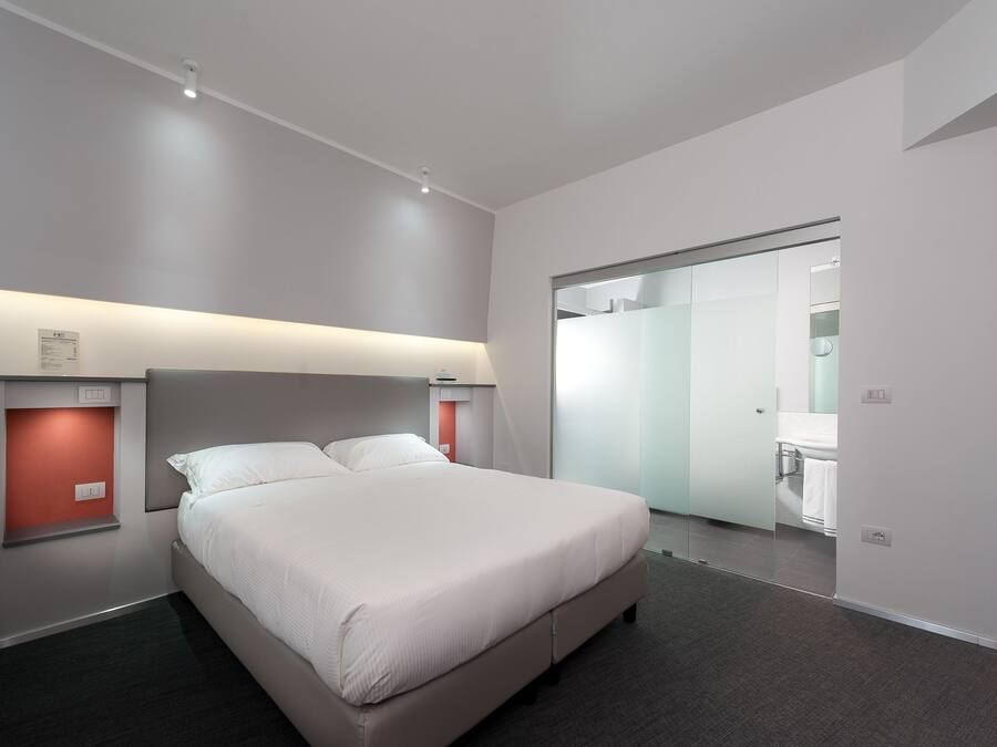 Una habitación de hotel moderna con una cama grande, una mesita de noche, un espejo y una puerta corredera que da al baño.