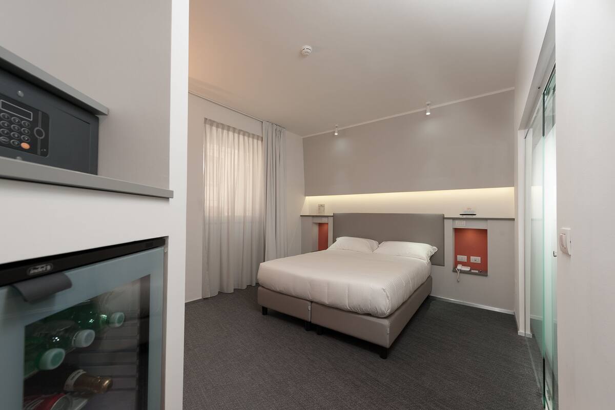 A modern hotel room with a bed, bedside tables, a minibar, and a glass partition wall.