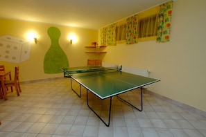 Una mesa de ping pong en una habitación con paredes amarillas, un adorno verde en la pared y una ventana con cortinas estampadas.