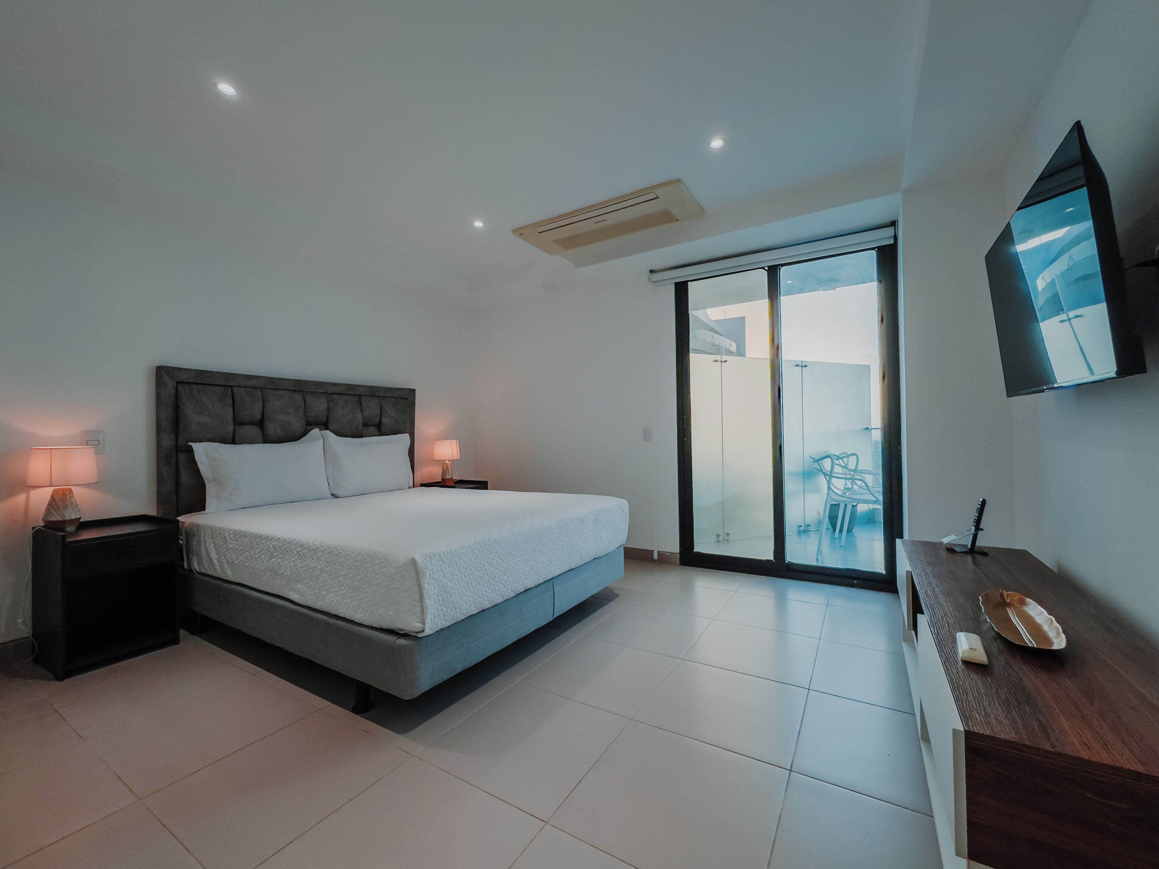 A modern bedroom with a large bed, bedside lamps, a TV, and a sliding glass door to a balcony.