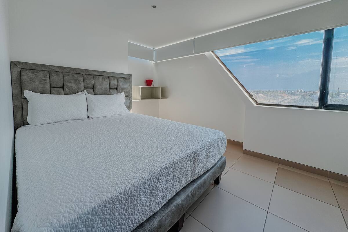 Une chambre avec un grand lit, une tête de lit grise, un puits de lumière et une vue sur la ville.