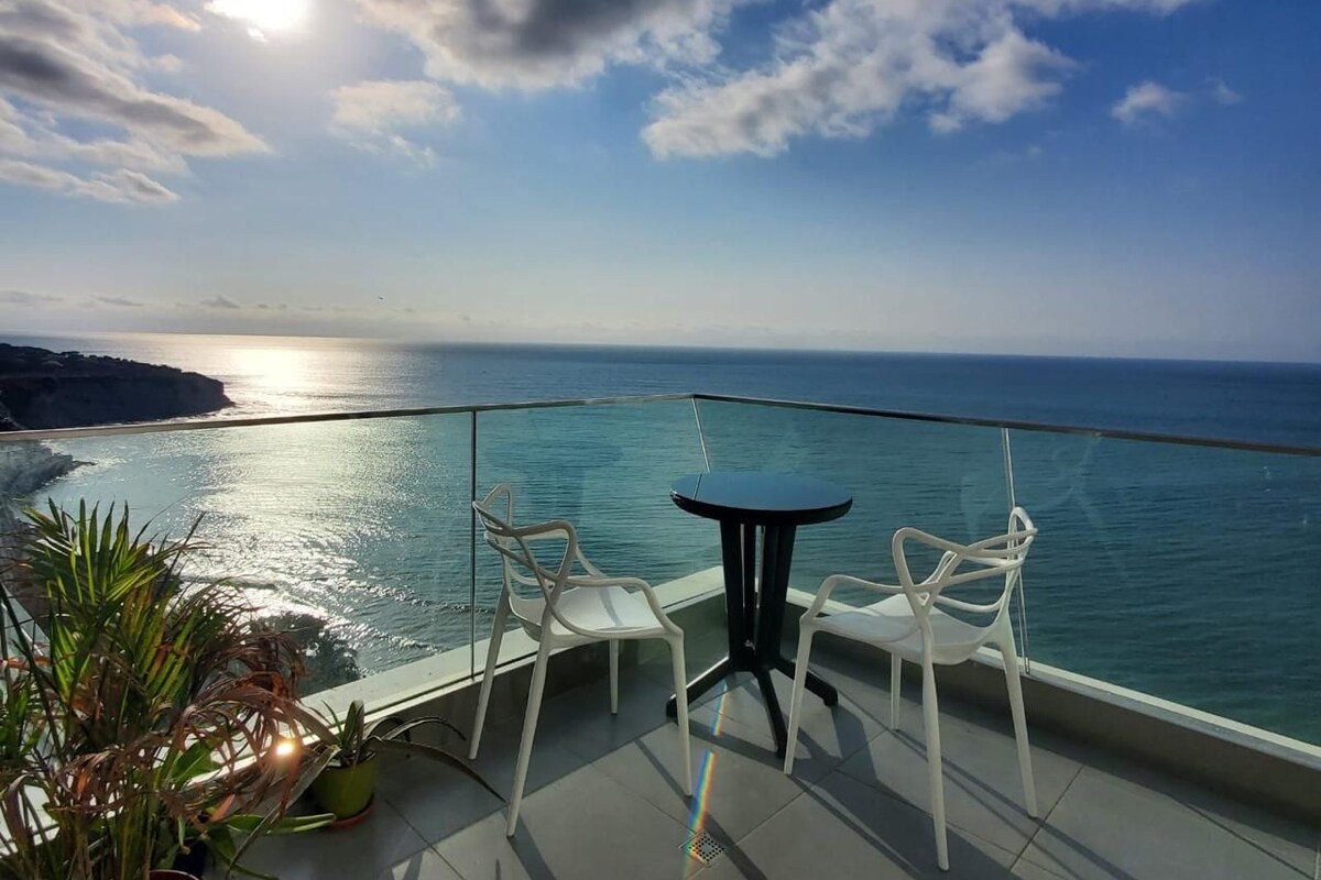 A balcony with a glass railing overlooking a coastal view, featuring two white chairs and a small round table.