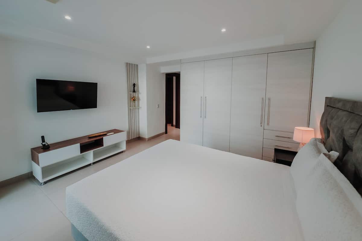 Une chambre moderne avec un grand lit, un téléviseur fixé au mur, une armoire et une table de chevet avec une lampe.