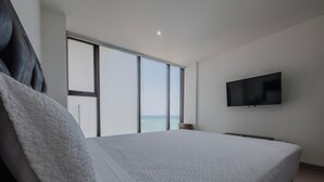 A modern bedroom with a large bed, a television mounted on the wall, and a view of the ocean through large windows.