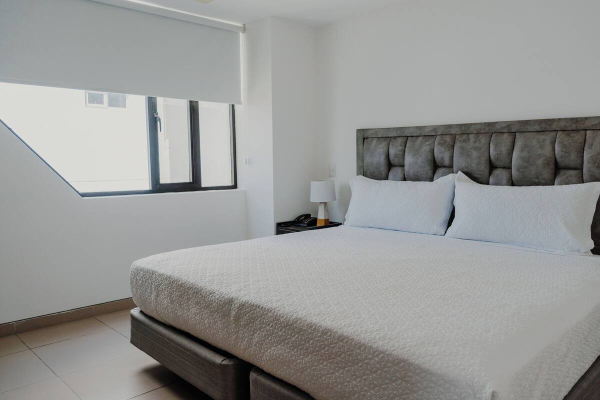 Une chambre d’hôtel moderne avec un grand lit, une tête de lit, une table de chevet avec une lampe et une fenêtre donnant sur l’extérieur.