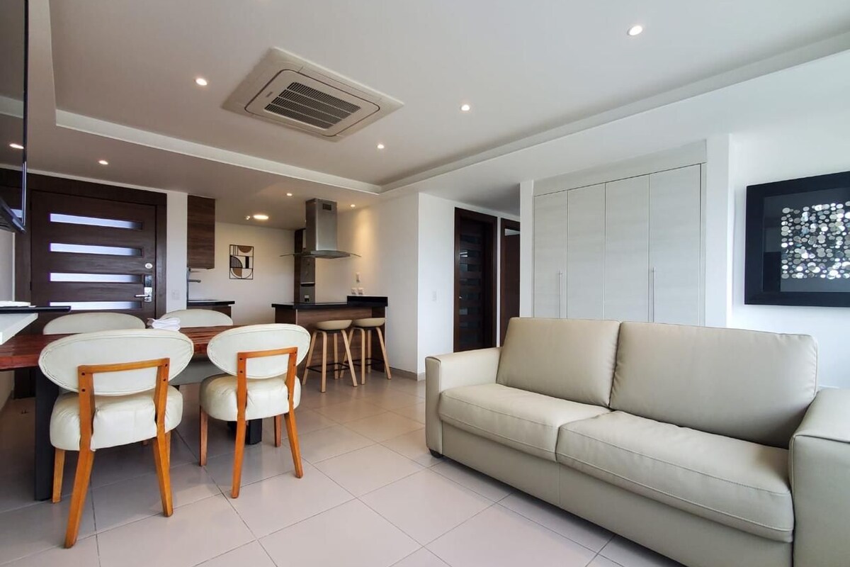 A modern living room with a beige sofa, a dining table with chairs, and a kitchen area in the background.