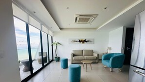 A spacious living room with a large window overlooking the beach, a beige sofa, a blue armchair, and a kitchen area with a refrigerator.