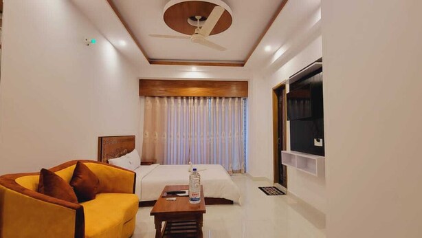Habitación de hotel con cama, un sillón amarillo, una mesita de noche de madera, un ventilador de techo y un gran ventanal con cortinas.