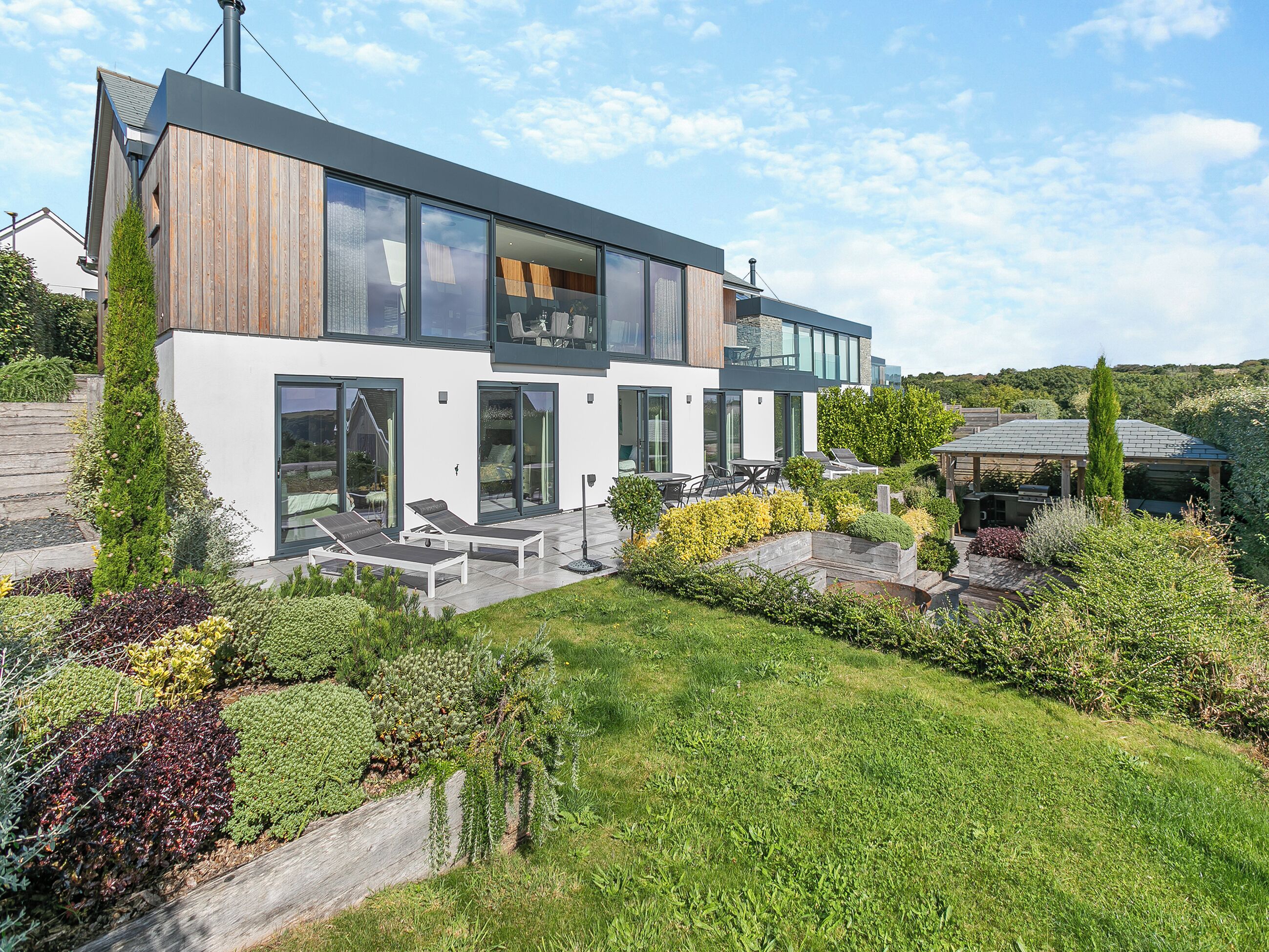 A modern two-story house with a large glass facade, a wooden deck, and a well-maintained garden.