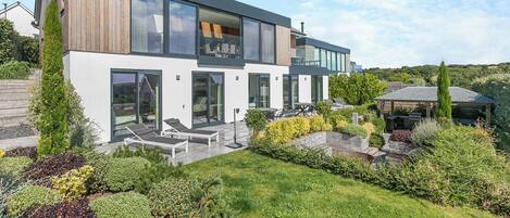 A modern two-story house with a large glass facade, a wooden deck, and a well-maintained garden.