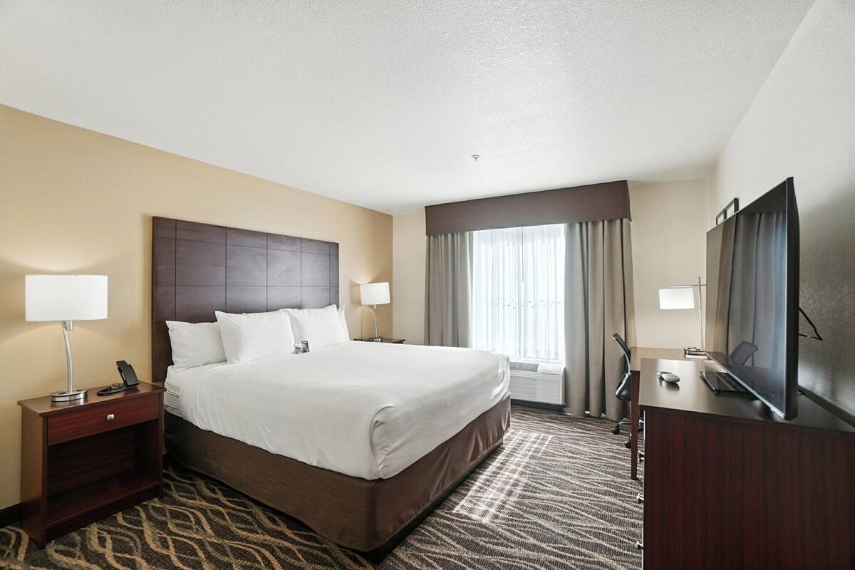 Quarto de hotel com uma cama grande, duas mesas de cabeceira, uma escrivaninha com cadeira, televisão e janela com cortinas.