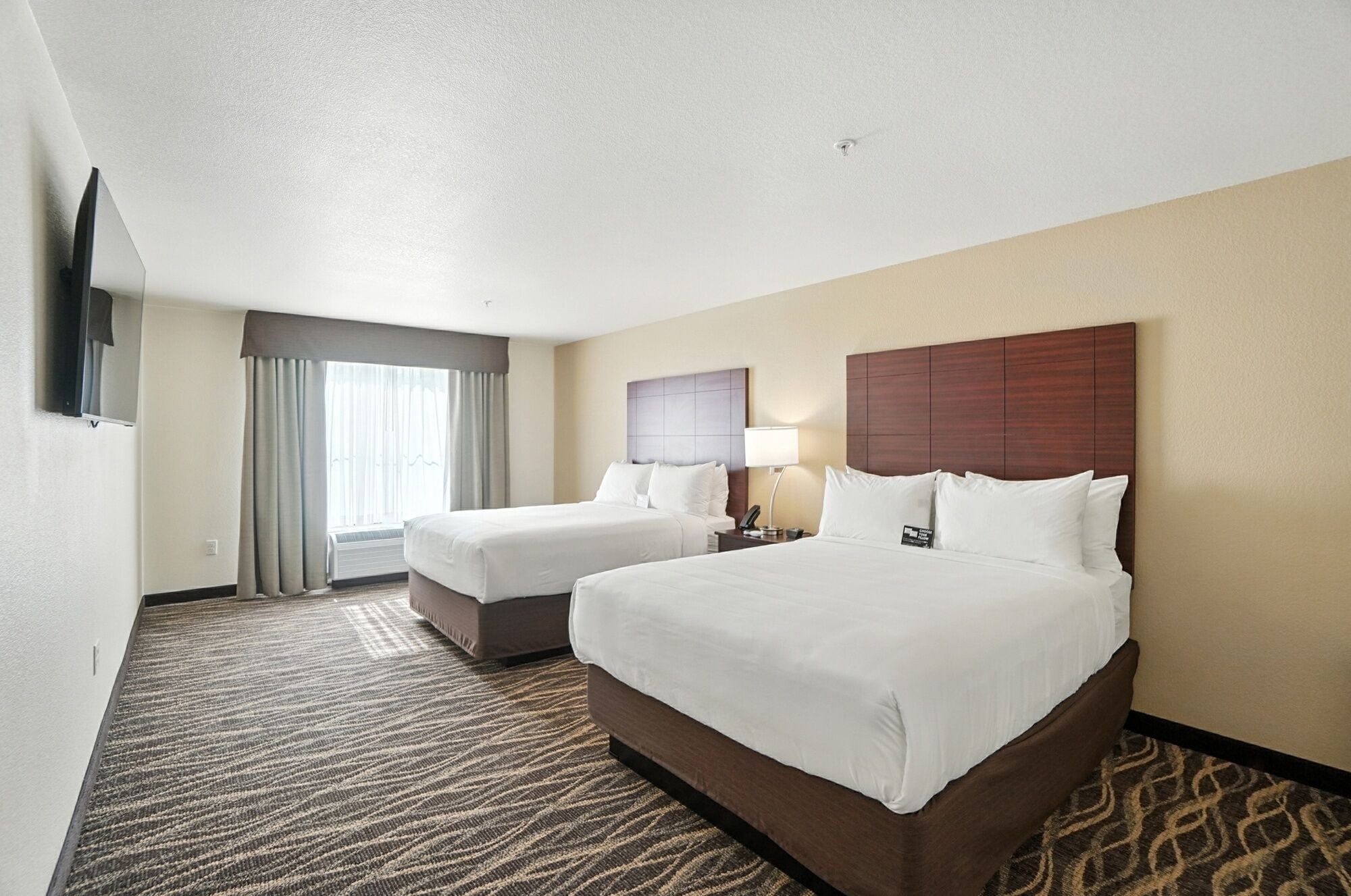 A hotel room with two beds, a flat-screen TV, a desk with a lamp, and a large window with curtains.