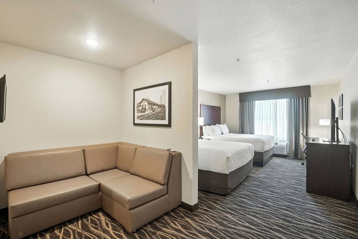 Quarto de hotel com sofá bege, duas camas, uma escrivaninha e um quadro na parede.