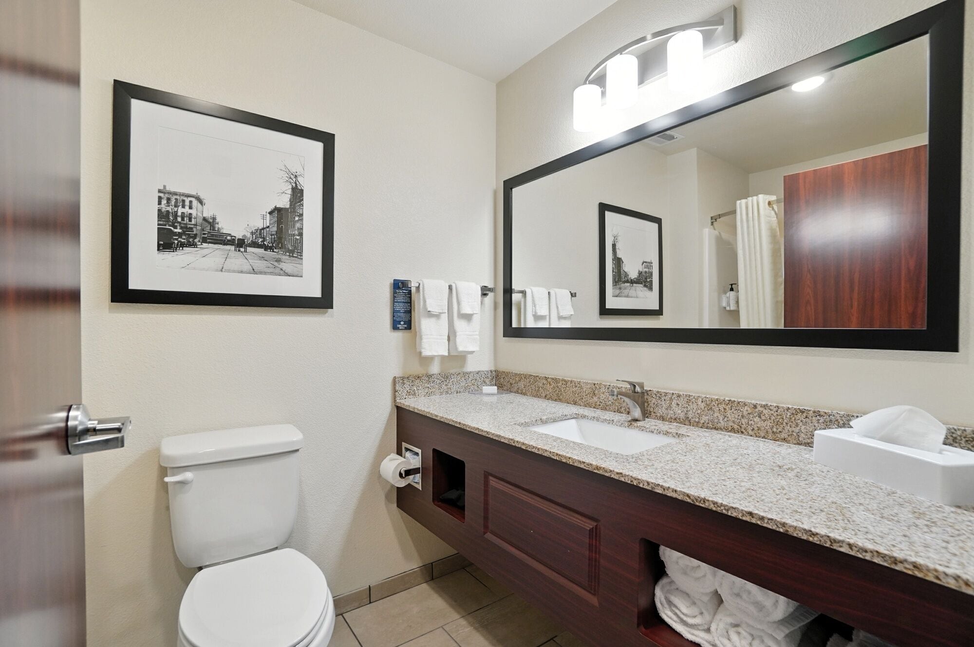 A bathroom with a toilet, sink, and large mirror. There are towels hanging on the wall and a framed picture above the mirror.