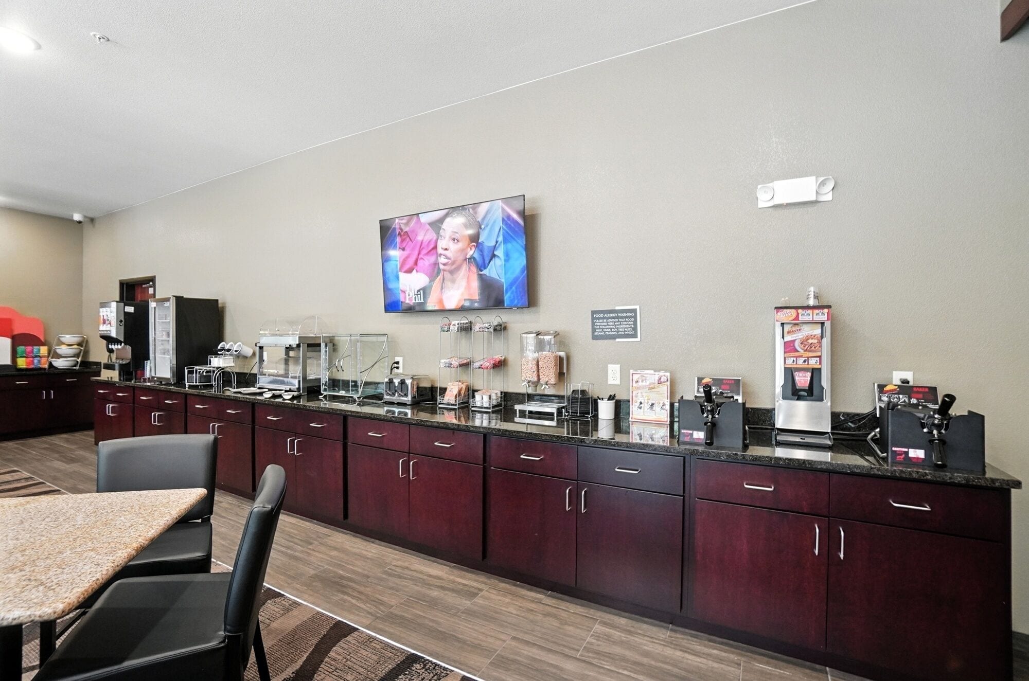 Un vestíbulo de hotel con una larga recepción, un televisor en la pared y diversas comodidades como una máquina de café y dispensador de zumo.