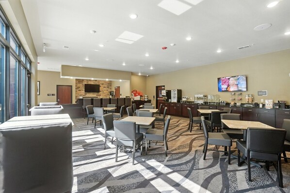 Un comedor de hotel con varias mesas y sillas, una zona de cocina y un televisor en la pared.