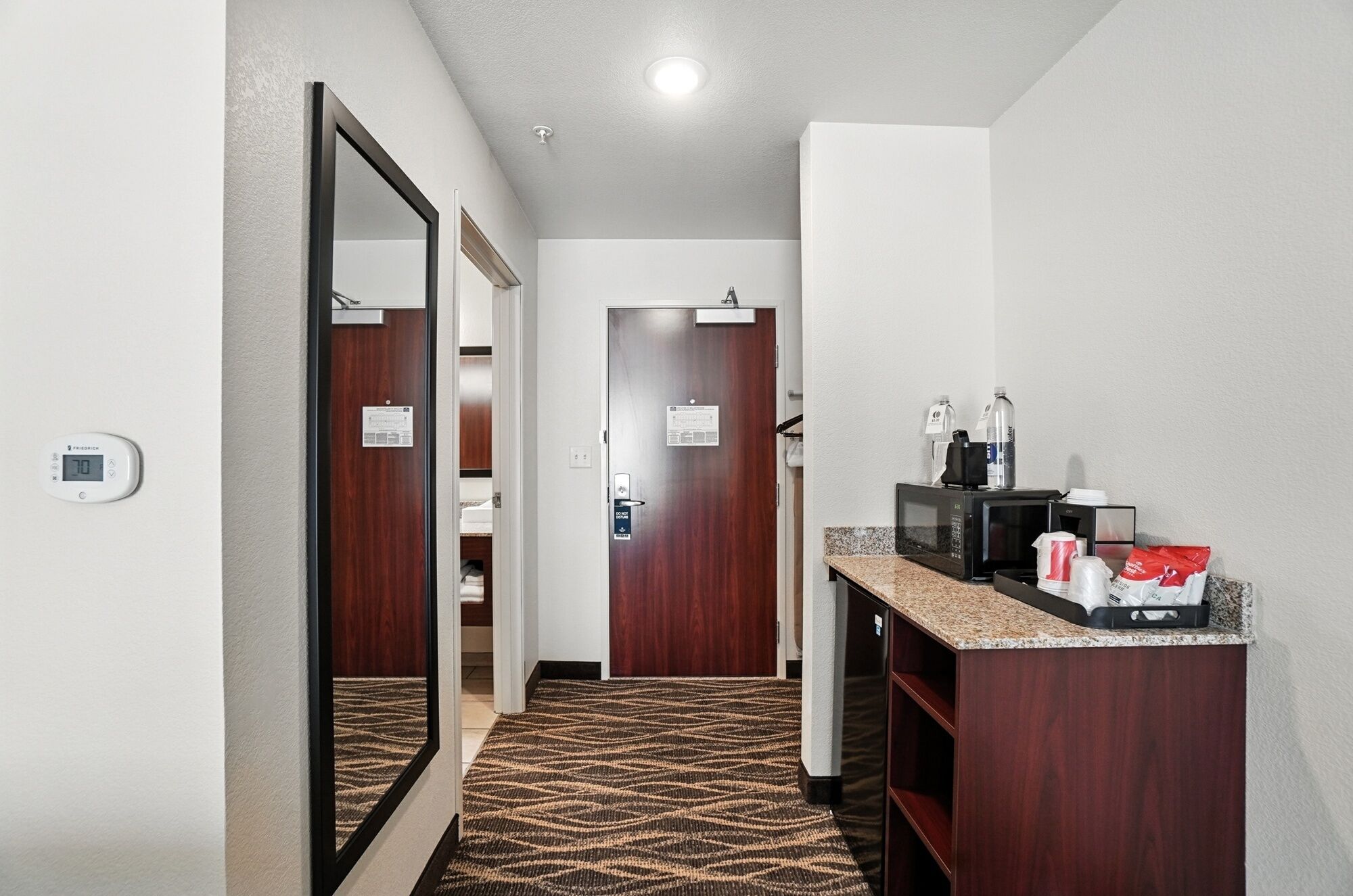 A hotel hallway with a carpeted floor, a mirror, a microwave, and a small counter with items on it.