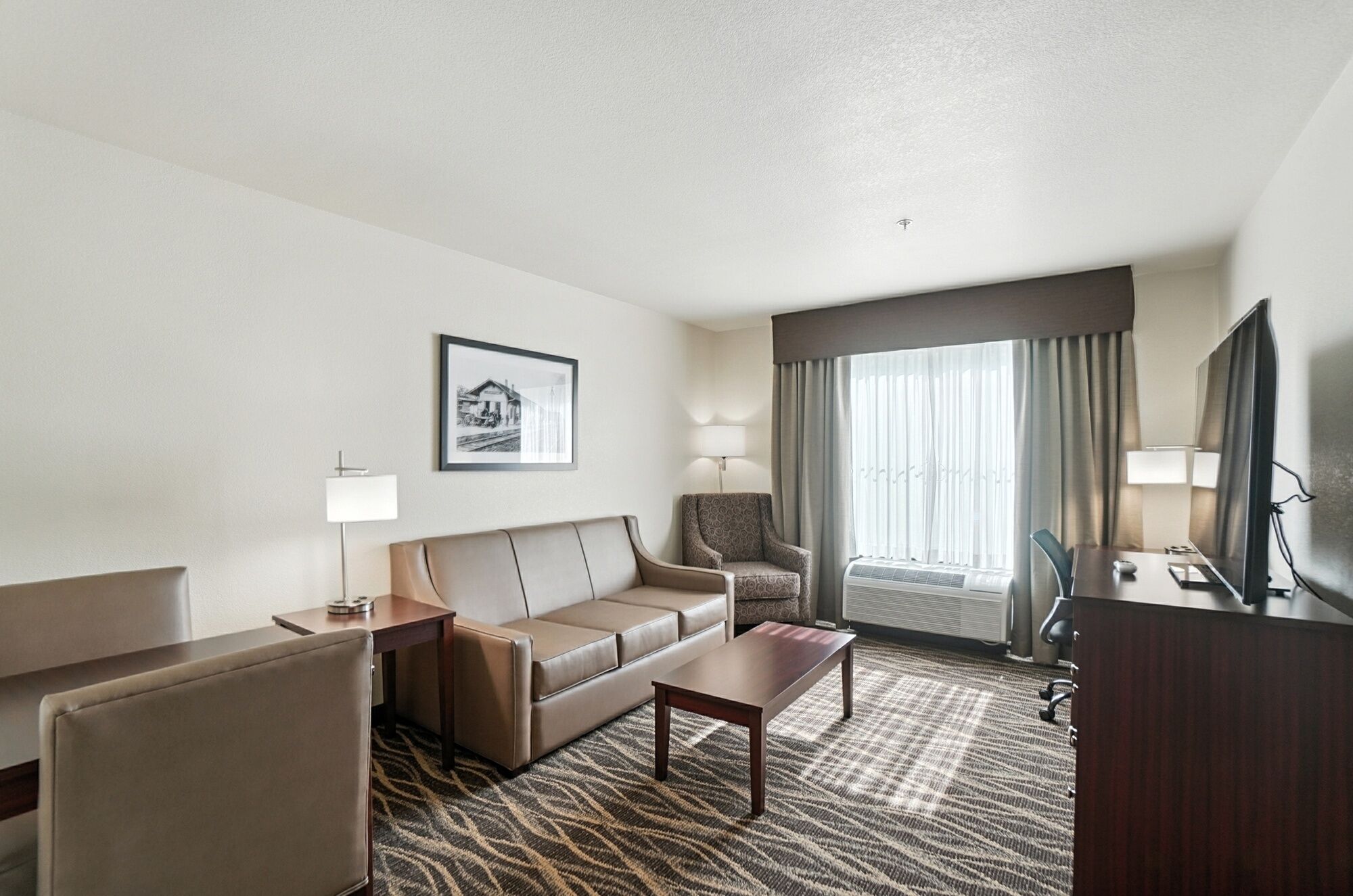 A hotel room with a sofa, a small table, a chair, a desk, a television, and a window with curtains.